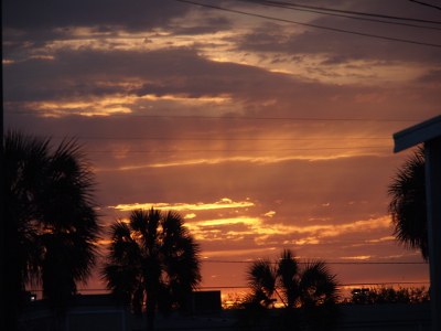 [The rays of the sun are visible above the trees and buildings, but the sun has not yet cleared the horizon. The trees are palms and the sky which is a blanket a clouds has colors ranging from yellow-white to purple-blue.]